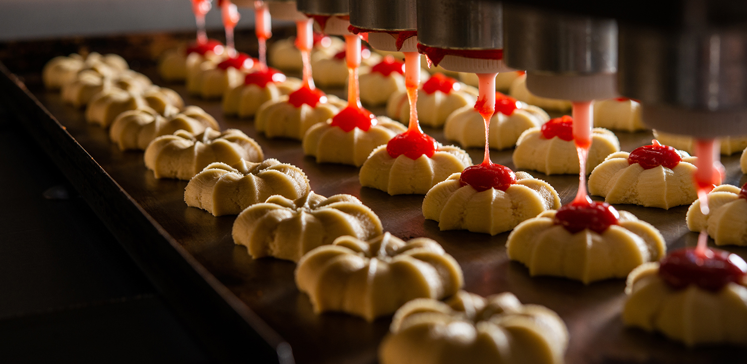 Shortbread cookies being filled with red filling on a conveyor belt.